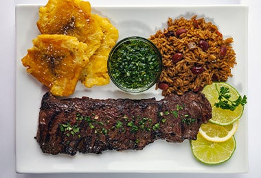 Un plato con chimichurri, carne, arroz y patacones.