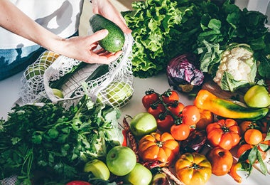 Verduras y frutas organizadas sobre una mesada blanca y una persona sacando otras de una malla blanca