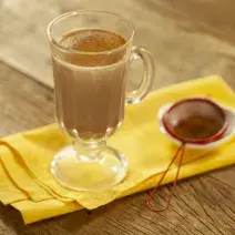 Fotografia em tons de marrom e amarelo de uma bancada de madeira com um paninho amarelo, sobre ele uma xícara transparente com leite, chocolate baton derretido e canela em pó. Ao lado uma peneira vermelha com canela.