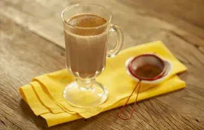 Fotografia em tons de marrom e amarelo de uma bancada de madeira com um paninho amarelo, sobre ele uma xícara transparente com leite, chocolate baton derretido e canela em pó. Ao lado uma peneira vermelha com canela. Fotografia em tons de marrom e amarelo de uma bancada de madeira com um paninho amarelo, sobre ele uma xícara transparente com leite, chocolate baton derretido e canela em pó. Ao lado uma peneira vermelha com canela.