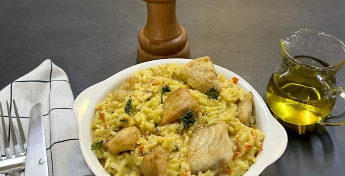 Foto da receita de arroz com peixe cremoso servida em uma panelinha de porcelana branca, ao lado esquerdo há talheres prateados sobre um pano quadriculado branco e preto. Ao fundo, um dispenser de azeite de vidro e um moedor de sal